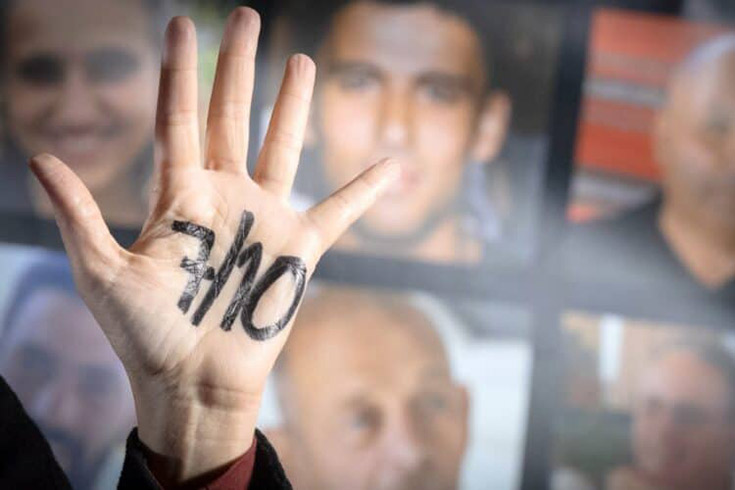 A hand, palm facing forward, with 7/10 displayed on it in thick, black ink, against a backdrop of blurred images of the faces of hostages