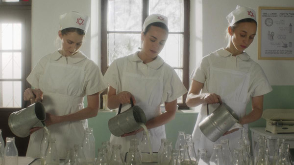 Three women in nurses' uniforms pour milk from metal pitchers into glass bottles.