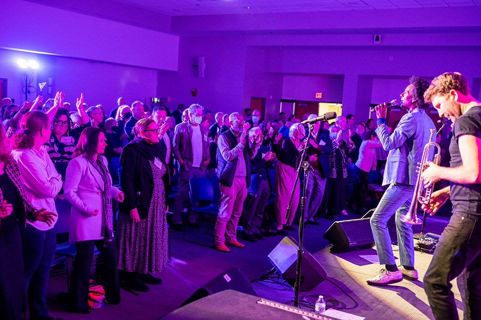 A concert audience standing and clapping while a man sings onstage and another man plays the trumpet