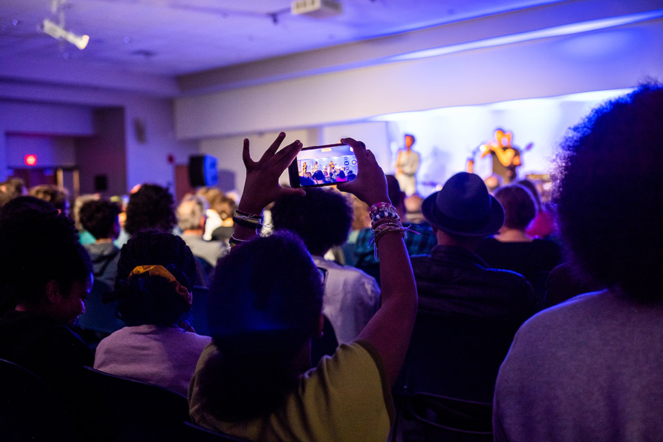 Woman in a large concert audience holds up smart phone photographing or video-recording the performers on the stage