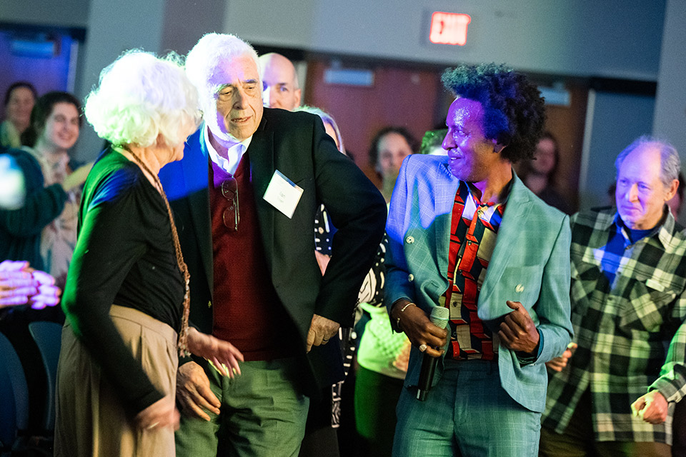 Man in blue suit dances next to man in dark suit amidst dancing people