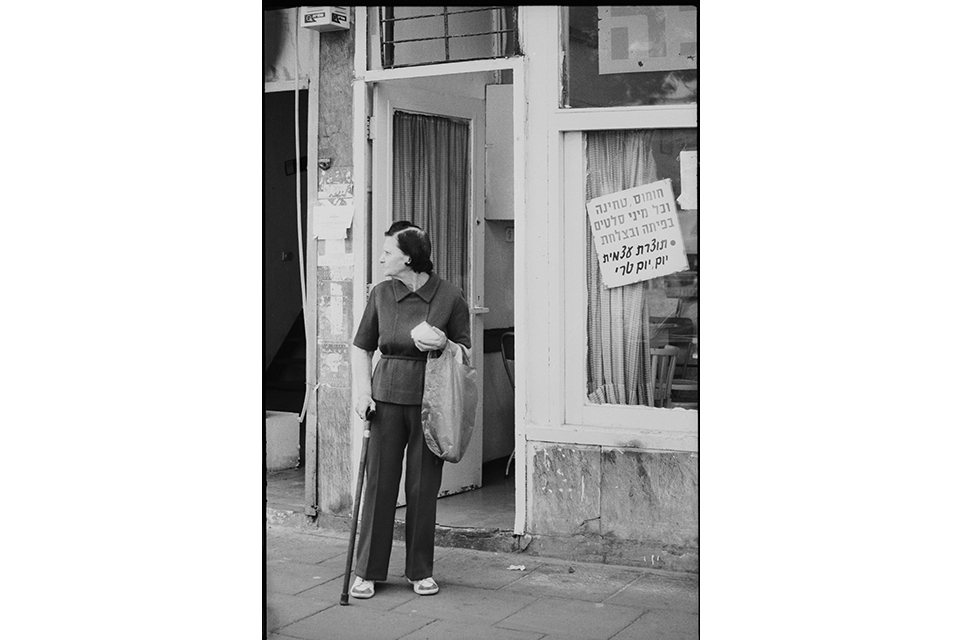 Woman standing on street outside a shop, with a cane, and a shopping bag on her arm