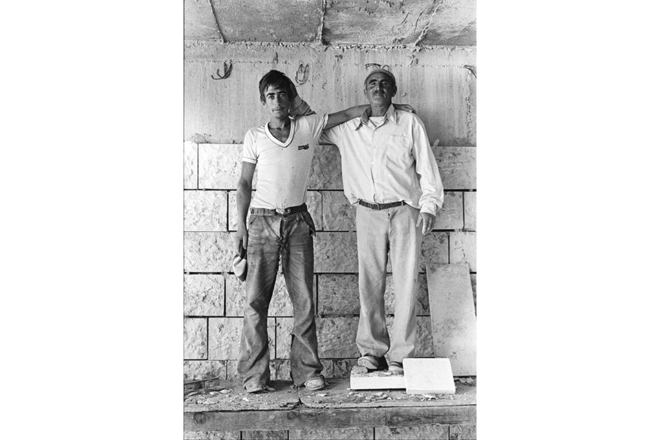 Two men standing side by side. in front of a partially built stone wall