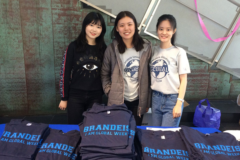 Three students wearing shirts saying I am Global Week shirts stand behind a table.