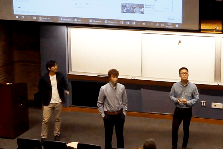 three students on stage presenting their work in front of projection screen