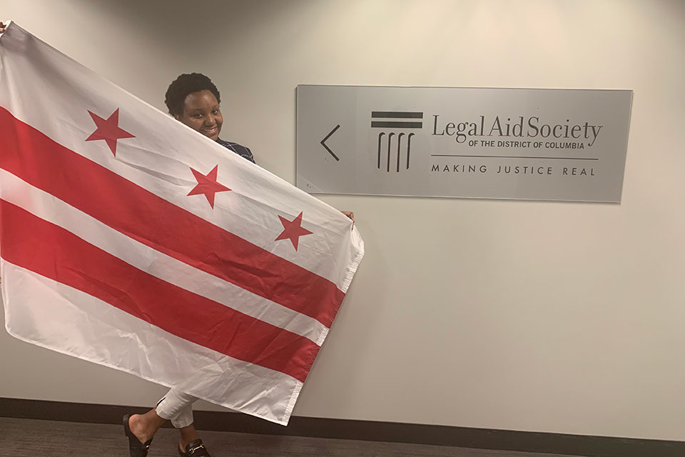 A student stands holding the flag of Washington D.C. in front of a sign that says Legal Aid Society of the District of Columbia: Making Justice Real