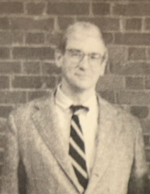 Professor Lyman Stookey, a man wearing a suit jacket, striped tie, and black glasses.