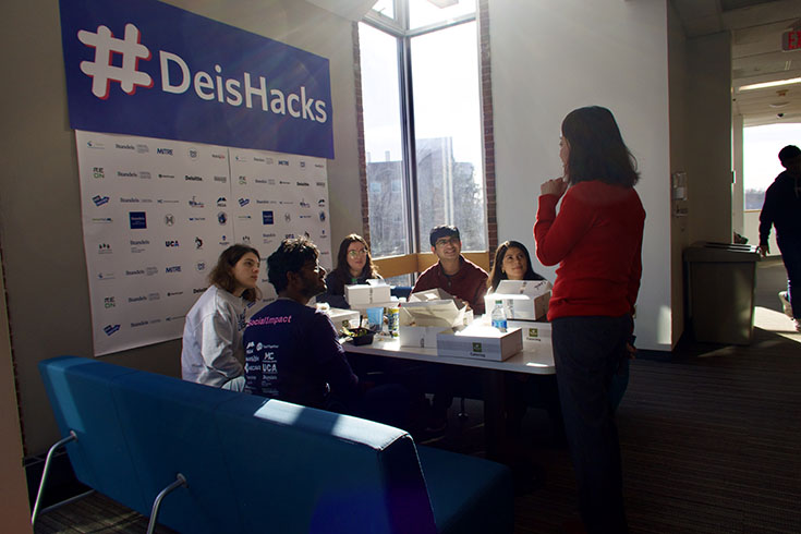 A group of students sit around a table and listen to a mentor. They sit in front of a large blue sign reading "DeisHacks".