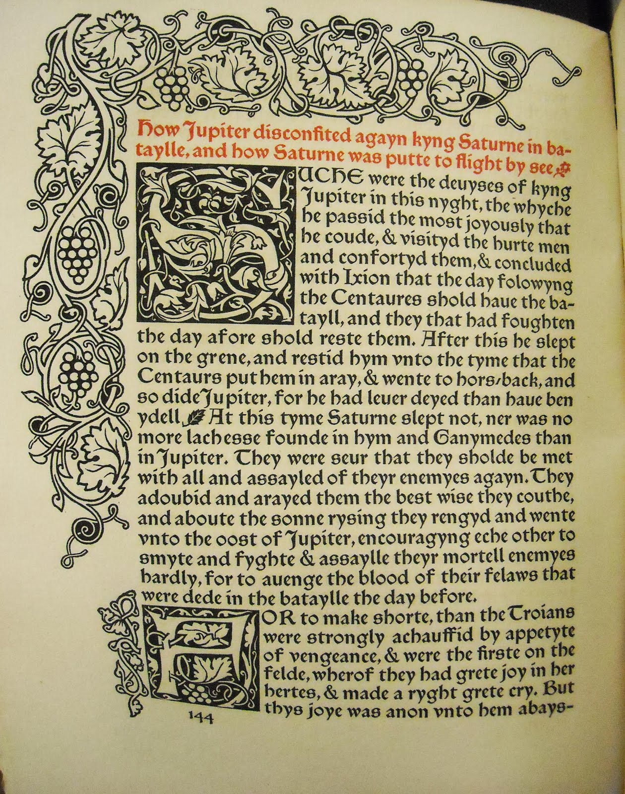 A page of The Recuyell of the Historyes of Troye with ornate vine and fruit border, two illuminated letters and orange first sentence.
