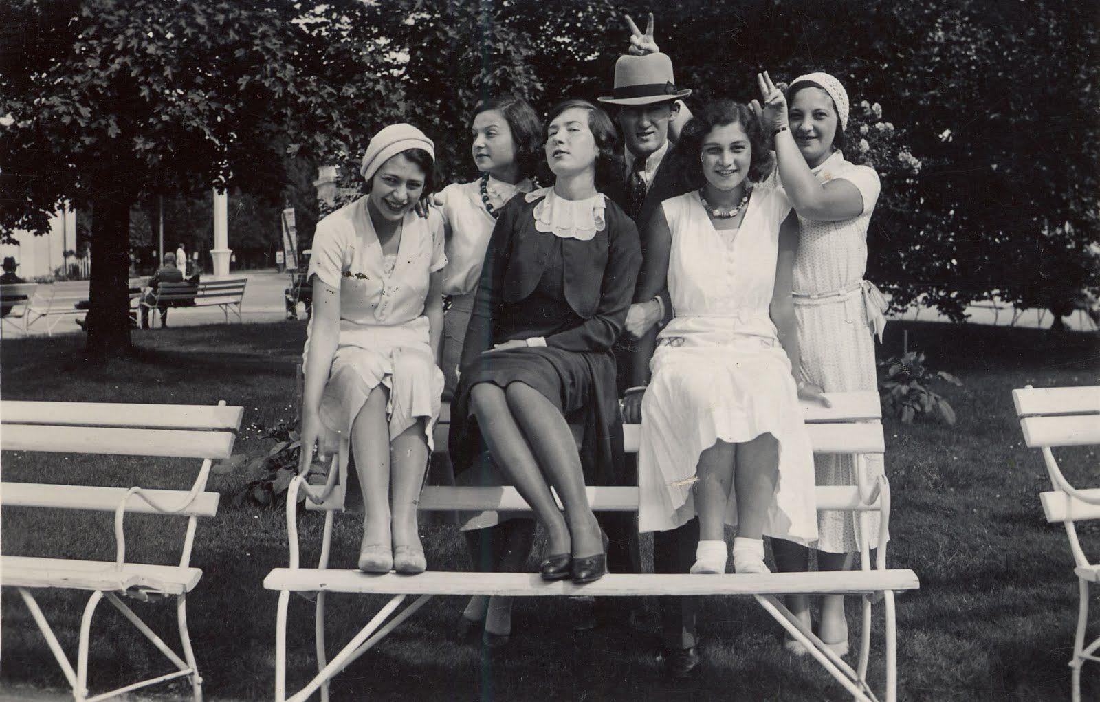 Family photograph of 5 women and one man at a park bench. The woman on the right holds up two fingers behind each of the two people next to her to make donkey ears. 