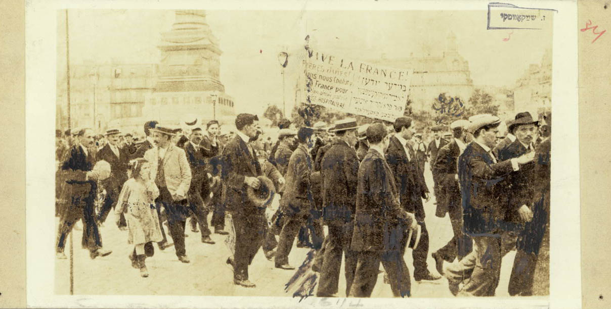 Photograph of a rally, with men and children marching, some holding a large sign written in French and Yiddish. The largest letter say "Vive la France"