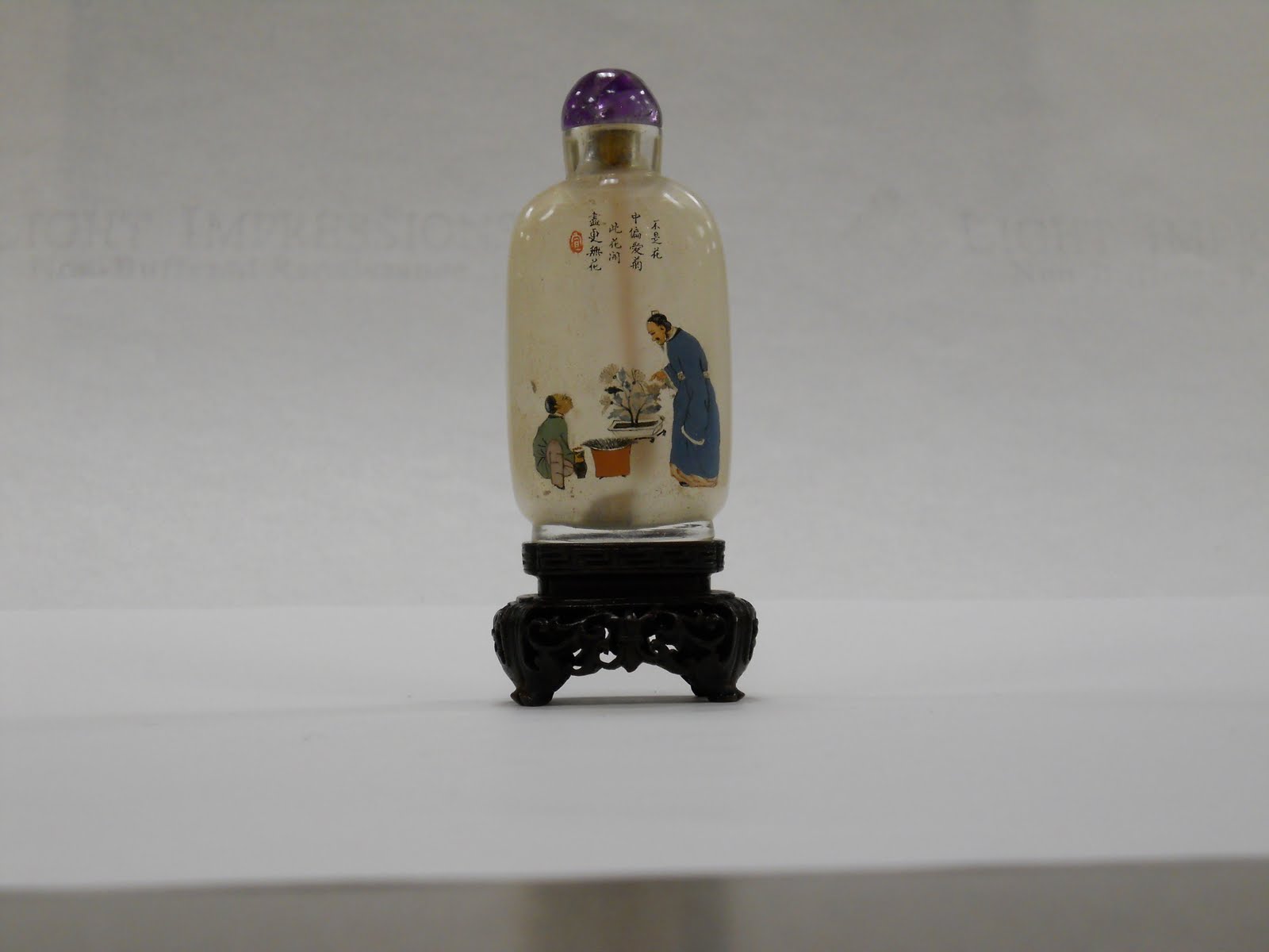 Chinese snuff bottle with black Chinese characters above a colorful illustration of an older gentleman who is tending to a small tree in the presence of a younger male figure. The bottle is predominantly beige and has a deep purple top.