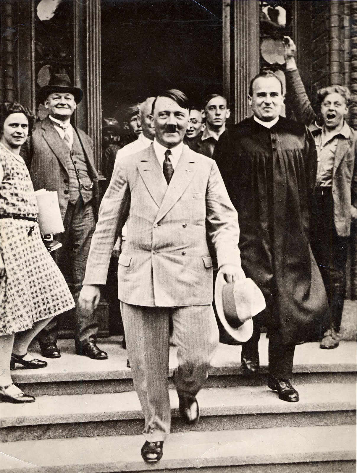 Adolf Hitler, wearing  a suit, walking down 3 steps outside of a building with a crowd of people behind him.