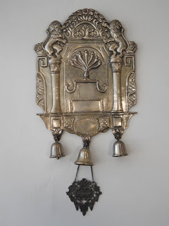 Silver ritual object hanging on a wall with 3 small bells. The low relief designs in the silver include 2 lions facing each other, each perched on top of a column.  In the center of the piece is a menorah.