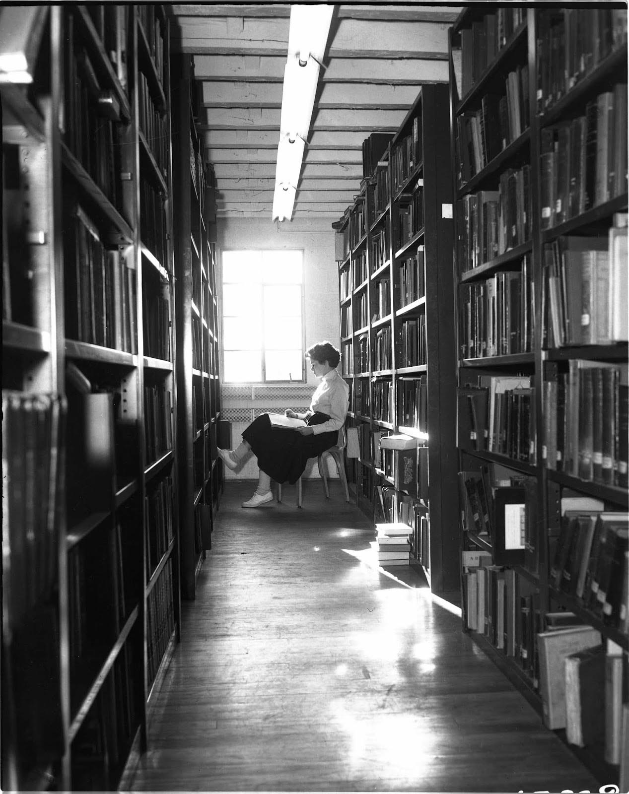 Black and white photo of the library stacks with a student sitting at the end.