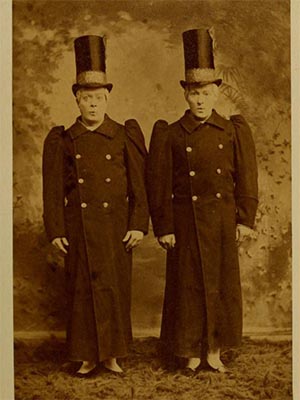 Sepia photograph of two actors wearing matching long coats, wigs and tall top hats