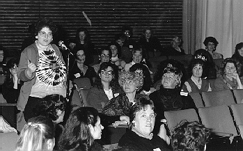 Tenth Anniversary of Women's Studies Program. A woman wearing a tied dyed shirt stands at the mic in the aisle of an auditorium filled with women