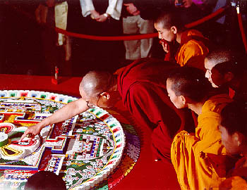 His Holiness the Dalai Lama and Buddhist Nuns dismantling the Mandala May 1998. Buddhist nuns in orange robes stand on either side of him.  People watch from behind a velvet rope barrier.