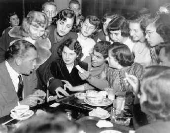 Ed Sullivan Visit. Ed Sullivan is seated at a table surrounded by a thick crowd of students.