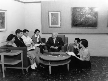 Students with Dr. Ludwig Lewisohn, J.M. Kaplan Professor Comparative Literature. Four students are seated on a sofa to Dr. Lewisohn's right. Two students kneel on the floor to his left.