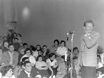 Danny Kaye standing at a mic looking away from the audience, smiling.  The students in the audience close to Kaye are seated on the floor close together. Behind them are students sitting on chairs. More students are seated on the floor in the aisles. Everyone is smiling.