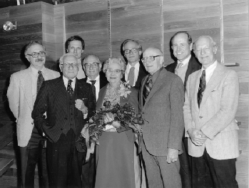 Gertrude Carnovsky, Assistant Dean of Faculty Retirement party after 28 years. Group photo of Carnovsky in the center holding a bouquet of flowers, surrounded by eight men wearing suits and ties.
