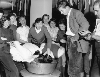 Students at a party.  Two students are dunking their heads in a basin of water while another student who is looking at the camera has his hand on the head of one of these students. Many others are standing around watching.  
