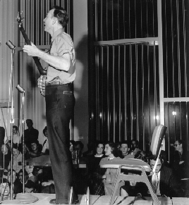 Pete Seeger standing on a stage playing a banjo and singing before an audience of students.