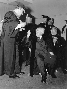 Israeli Prime Minister Ben-Gurion at Convocation. He is seated, wearing academic regalia as are the other people nearby.  One man is standing next to him talking with him.