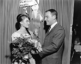Photo of a woman and man in formal dress facing each other at a party in front of a sign that says "Playboy Party with a cutout of a large cartoon bunny holding a champagne glass." The woman holds a bouquet.