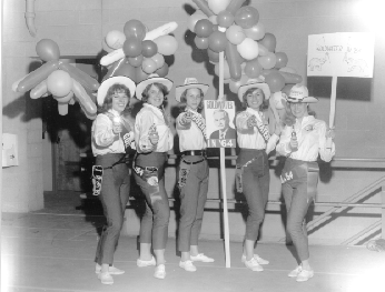 Five women wearing matching cowboy hats, holsters, white blouses standing in a row with a campaign poster for Barry Goldwater. Behind them are clusters of balloons.
