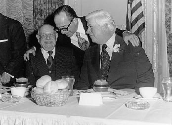 Congressman Tip O'Neil at a dinner at Copley, November 16, 1975. A man stands behind him and places a hand on his shoulder and the shoulder of the man seated next to O'Neil.