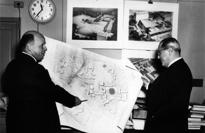 Two men looking at Brandeis Plans. The man on the left holds a very large piece of paper with the map of Brandeis in one hand, and his other hand holds a pencil which he is pointing at a spot on the map.