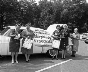 Five women, holding books and shopping bags,  standing in front of the Bookmobile, a car with the trunk propped open and large banner on the side of the car that says: Brandeis Women's Committee Mobile Book Pickup [word hidden]. New books for Old [word hidden].