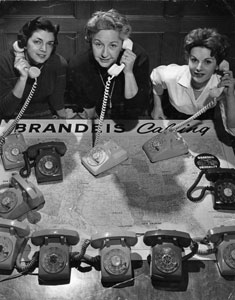 Three women, sitting at a table with 11 rotary phones in front of them, each holding a phone receiver to their ears.