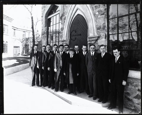 Group of 12 men wearing suits and ties standing in front of Castle. In the center is an older man wearing a hat and an overcoat.