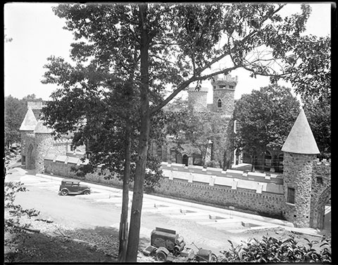 View of the Castle behind a sonte wall with two archways that have a little cylindrical tower and pointy roof on either side.  A car is parked by the wall.