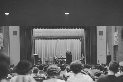 Meeting held at Seifer Auditorium. Four people are on the stage facing a filled auditorium.