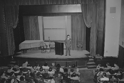 Max Lerner stands on the stage next to the lectern addressing a full auditorium.