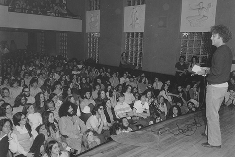 Abbie Hoffman '59 on the Nathan Seifer Auditorium Stage addressing an audience that fills the room including many who are standing in the aisles.
