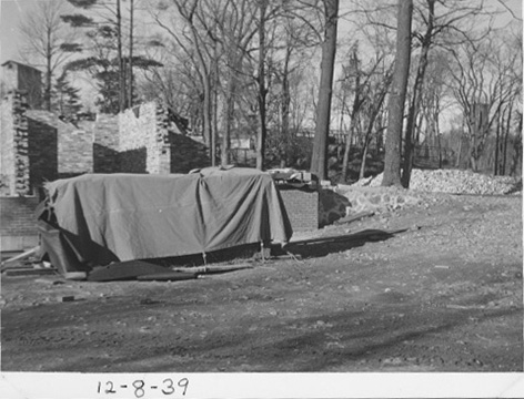 A pile of supplies covered by a tarp, on the ground in front of the partially built brick walls.