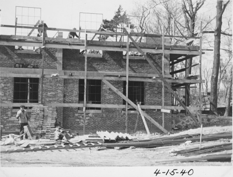 Work crew are installing windows. 2 men stand on the top level of the building while one works on the ground below.