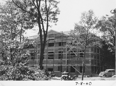 Close to completion, this photo shows the building with its top floor and roof.  There are cars parked out front and several trees partially obscure the view.