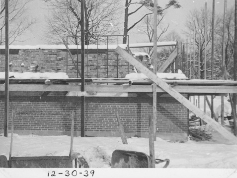 A window frame being installed in the brick wall. Wood posts and scaffolding are in front of the partially built brick wall.