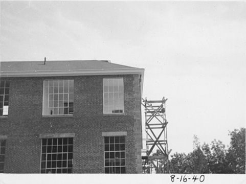 Photo of nearly completed building, with roof and windows, and minimal scaffolding on one side.