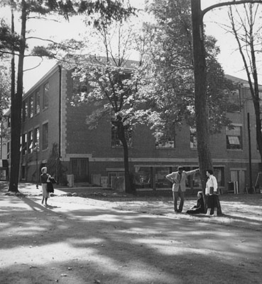 Back of Ford Hall. October 9, 1953. The building is visible through a few trees. Three students talk in front of a tree. Another student walks down the path.