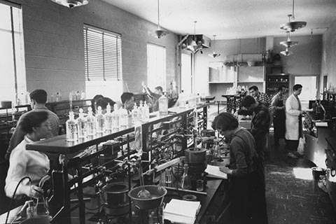 Students working in the Science Laboratory in Sydeman Hall, with a long narrow table in the center, lots of beakers, burners.  Teacher is wearing a lab coat.