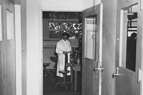 An open door reveals a science lab where a man in a lab coat is reading a paper he holds in his hands.