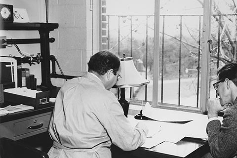 A view of a faculty office.  A man in a lab coat sits at a desk accompanied by another man in a suit.  Both men, seen from behind,  are looking at papers on the desk.