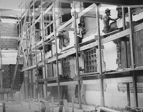 Three men perched on scaffolding, using hammers, while one stands below working on a window.